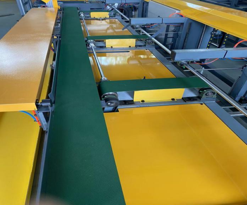 Feeding section of a profile packaging machine, showing a green conveyor belt flanked by yellow side panels and metal guide rails.
