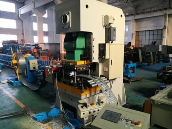 A large, off-white punch press machine is shown in operation within a factory, actively punching holes into a metal strip that is being fed through it as part of the Fully Automatic Cable Tray Production Line.