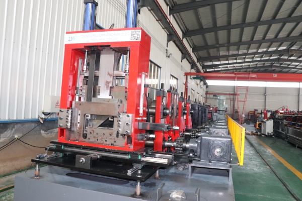 A large, red and silver CZ purlin making machine is displayed in a factory setting. The machine features a prominent red punching section at the front and a long series of forming rollers extending into the background. An overhead crane is visible above the machine.