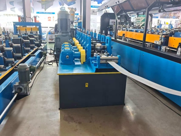A Split set roll forming machine in an exhibition hall, processing a long, flat metal strip. The machine is painted blue and features a series of rollers and motors.
