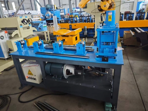 A Split set necking machine, painted blue and grey, is shown in a factory. It features a motor, a control box, and a series of rollers on its work surface, with finished metal rods lying on the floor. This machine is used for manufacturing Rock Bolts.