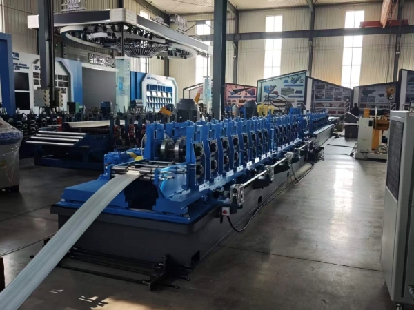 A long, blue CZ purlin making machine in a factory, processing a flat metal strip fed from the foreground. The machine features a series of roller dies along its length.