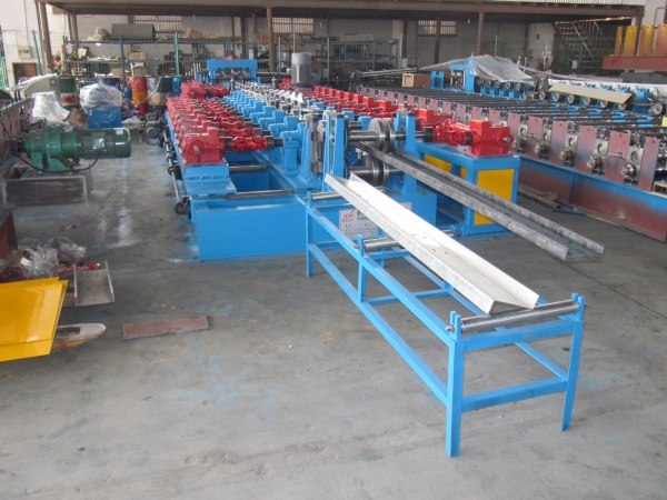 A long, blue and red CZ purlin making machine is shown in a factory, with a finished white purlin resting on a blue support rack in the foreground.