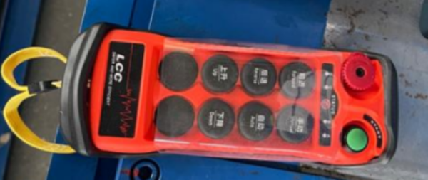 An orange and black industrial wireless remote controller with multiple black function buttons, a green start button, and a red emergency stop knob, shown resting on a blue surface.