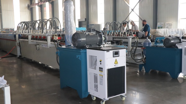 T-Bar Roll Forming Machine on display in a factory, showing the main production line with its motor, hydraulic system, and a separate white cooling unit.