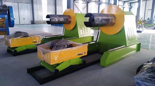 Two green Hydraulic De-Coiler units with yellow coil supports and black mandrels, positioned side by side in a factory setting; some components are wrapped in protective plastic.