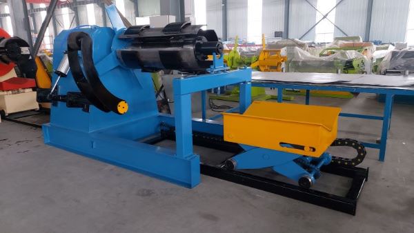A 7-ton hydraulic de-coiler machine in a factory setting, featuring a blue-painted steel frame, a large black coil-holding mandrel with hydraulic arms, and a yellow-colored support tray on a scissor-lift mechanism, positioned next to a metal worktable.